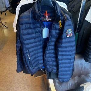 Mörkblå dunjacka Parajumpers XXL - Snygg mörkblå dunjacka från Parajumpers i storlek XXL. Jackan har quiltad design, dragkedja framtill och coolt PJS-märke på ärmen. Insidan är blå och jackan har en gul detalj vid kragen. Perfekt för dig som vill ha en lätt och stilren jacka med streetkänsla.