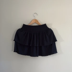 Svart volangkjol från Kappahl - Supersöt svart kjol från Kappahl med två lager volanger och smockad midja. Kjolen är kort och materialet är tunt.