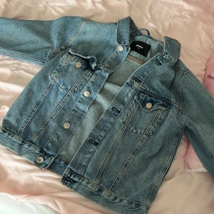 Klassisk jeansjacka från Bik Bok XS - Snygg ljusblå jeansjacka från Bik Bok i storlek XS. Jackan har klassisk design med silverfärgade knappar, två bröstfickor med lock och raka sömmar. Perfekt att slänga över en hoodie eller t-shirt för en chill look.