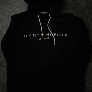 Svart hoodie Tommy Hilfiger S - Svart hoodie från Tommy Hilfiger i storlek S med vit snörning och klassisk logga broderad på bröstet. Tröjan har huva, känguruficka och långa ärmar. Perfekt för chill dagar och streetwear-stil. Materialet är mjuk bomull för skön komfort.