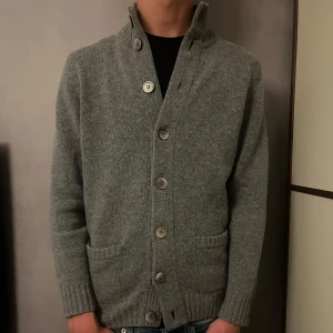 Alan Paine cardigan - Snygg grå kofta från Alan Paine med ribbad krage och stora knappar framtill. Tillverkad i mjuk ull, har två fickor och långärmad design. Perfekt för dig som gillar klassisk stil med modern touch. Passar dig som vill ha en varm och stilren tröja.