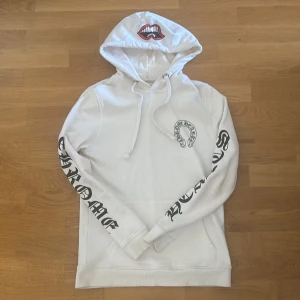 Vit hoodie Chrome Hearts S - Vit hoodie från Chrome Hearts med coolt tryck av röda läppar på huvan och svart logga på bröstet. Snygga detaljer med snörning och metallöglor. Svarta texttryck längs båda ärmarna ger en edgy vibe. Perfekt för dig som gillar streetwear och statement-plagg.