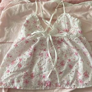 Blommig vit linnetopp från SHEIN - Supersöt vit linnetopp från SHEIN med rosa och ljusgula blommor, broderade detaljer och små volanger vid axelbanden. Toppen har smala axelband, knytning framtill och är lätt genombruten med broderade hål. Perfekt för en somrig look!
