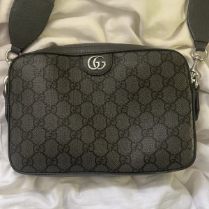 Svart axelväska från Gucci - Säljer en snygg svart axelväska från Gucci med klassiskt GG-mönster över hela väskan. Väskan har en bred, justerbar axelrem och detaljer i skinn. Perfekt storlek för det viktigaste och har en stilren, rektangulär form med silverfärgad logga framtill. Den kostar ny 17,300kr.