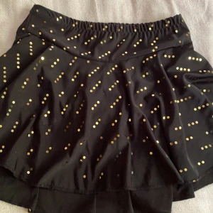 Svart kortkjol med guldprickar SkateJoy konståknings kjol❤️ - Supersnygg svart kortkjol från SkateJoy med glittriga guldprickar över hela tyget. Kjolen har en elastisk midja och är gjord i ett mjukt, syntetiskt material som ger bra rörelsefrihet. Perfekt för dig som gillar att sticka ut med lite extra bling!