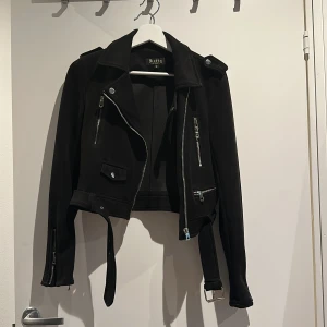 Svart bikerjacka från Softy Collection - Svart bikerjacka från Softy Collection i mocka-look med coola silverfärgade dragkedjor, bälte i midjan och detaljer vid axlarna. Croppad passform och långa ärmar med dragkedjor vid ärmslut. 