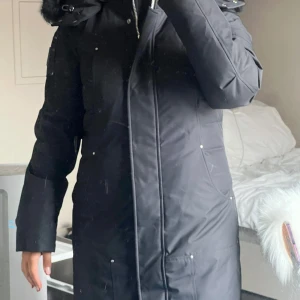 Svart dunjacka från Moose Knuckles - Svart Moose Knuckles jacka med dragkedja och knappar. Jackan har en huva med pälsdetaljer och flera fickor.  Köpte denna jacka förra sommaren och hann gå ner i vikt till vintern så jag fick ingen användning av denna. Vill sälja o köpa en ny