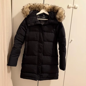 Svart lång dunjacka från Everest - Säljer en svart, lång dunjacka från Everest med fluffig fuskpäls på huvan. Jackan har quiltad design, dragkedja och knappar framtill. Perfekt för kalla vinterdagar och har en snygg, stilren look som funkar till alla outfits.