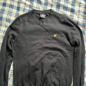 Svart stickad tröja från Lyle & Scott - Svart stickad tröja från Lyle & Scott med klassisk v-ringning och gul broderad logga på bröstet. Tröjan har långa ärmar och är gjord i mjukt material som passar perfekt för kyliga dagar. Enkel och stilren design som funkar till jeans eller chinos.