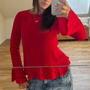 Röd långärmad blus med volang - Säljer en röd långärmad blus med snygga volangdetaljer vid ärmslut och nederkant. Toppen har öppen rygg med knyt och en cool layering-effekt. 