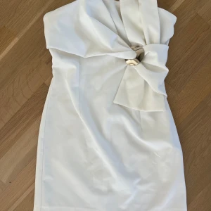 Vit axelbandslös kortklänning med rosett - Superfin vit axelbandslös kortklänning med stor rosett framtill och snygg metallring som detalj. Klänningen har en slim passform och är perfekt för dig som vill sticka ut med en elegant och modern look. Materialet känns mjukt och har en lätt stretch. Klänningen är ifrån Zara och har endast använts en gång. Säljer pga för klänningen inte kommer till användning.