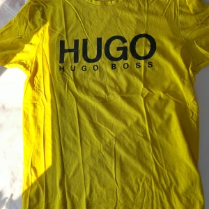 Gul t-shirt från Hugo Boss - Gul t-shirt från Hugo Boss med stor svart HUGO-logga på bröstet. Klassisk rund hals och korta ärmar. Tillverkad i mjuk bomull som känns skön mot huden.