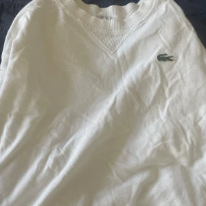 Vit piké Lacoste - Vit pikétröja från Lacoste med klassisk krokodillogga på bröstet. Tröjan har ribbad v-ringad krage och är tillverkad i bomull. Perfekt för dig som gillar clean och sportig stil. Långärmad modell som funkar året runt. Storlek L/M.