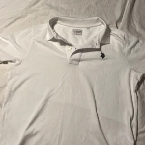 Vit pikétröja från U.S. Polo Assn. - Fräsch vit pikétröja från U.S. Polo Assn. med klassisk krage och korta ärmar. Tröjan har en liten broderad logga på bröstet och är gjord i mjuk bomull som känns skön mot huden. Perfekt för dig som gillar stilren och enkel design.