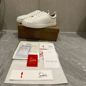 Christian Louboutin vita sneakers med nitar - Säljer ett par sjukt snygga vita sneakers från Christian Louboutin med ikoniska röda sulor och coola nitar på tån. Skorna har snörning och är tillverkade i skinn för en lyxig känsla. Perfekt för dig som vill sticka ut med en unik och trendig look.