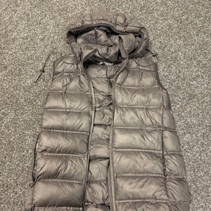 Svart dunväst från Zara med huva - Säljer en svart dunväst från Zara med quiltad design och avtagbar huva. Västen har dragkedja framtill och dragsko vid huvan för extra stilpoäng. Perfekt att slänga över en hoodie när det är kyligt ute.