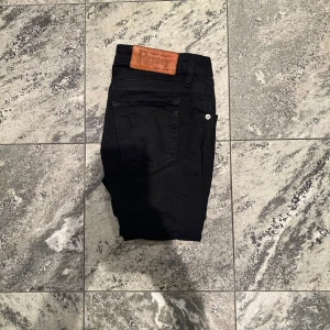 Svarta Replay jeans, storlek 25 - Säljer ett par svarta jeans från Replay i storlek 25. De har klassisk femficksdesign, snygga Replay-detaljer vid fickan och en brun läderpatch bak i midjan. Jeansen är raka och har en slim passform som sitter snyggt längs benen. Perfekta för dig som gillar stilrena och tidlösa jeans. 