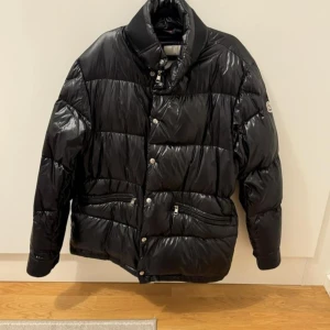 Svart Moncler pufferjacka herr - Svart pufferjacka från Moncler med glansig finish och klassisk logga på ärmen. Jackan har hög krage, dragkedja och knappar framtill samt två fickor med dragkedja. Insidan har en cool serietidningsetikett. Perfekt för kalla dagar och riktigt snygg streetstil.