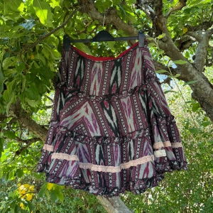 Mönstrad volangkjol med spetsdetaljer - Supersöt kortkjol från Odd Molly med färgstarkt mönster i svart, vinrött och grått. Kjolen har flera volanger och ljusrosa spetsdetaljer som ger en bohemisk vibe. Perfekt för dig som gillar unika plagg med mycket personlighet. Storlek 3 som enligt deras storleksguide motsvarar en storlek L.  Mycket fint skick, men kan ha någon enstaka lös tråd vid materiet vid midjan. Se sista bild. 