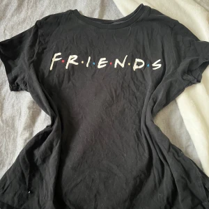 Svart Friends t-shirt med tryck - Svart t-shirt med klassiskt Friends-tryck på bröstet. Rund hals och korta ärmar, perfekt för dig som älskar serien. Mjuk och skön bomull som sitter snyggt och casual. Färgglada prickar mellan bokstäverna ger en extra touch.