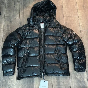 Svart pufferjacka från Moncler - Snygg svart pufferjacka från Moncler med glansig finish och huva. Jackan har quiltad design, dragkedja framtill och två sidofickor med dragkedja. Perfekt för kalla dagar när du vill hålla stilen. Moncler-logga på ärmen för extra premiumkänsla.