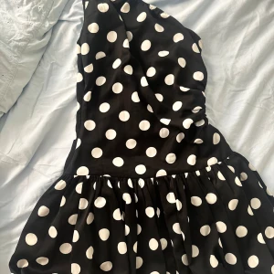 Svart prickig one shoulder klänning - Säljer en svart kortklänning med vita prickar och one shoulder-design. Kjolen är voluminös och har en snygg rynkad midja. Perfekt för dig som gillar att sticka ut med ett coolt mönster och unik form. Materialet känns lätt och luftigt.