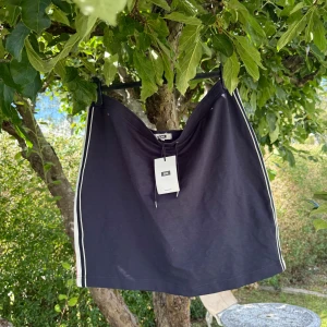 Svart kortkjol med snörning från WE - Sportig svart kortkjol från WE i storlek XXL. Kjolen har vita ränder längs sidorna och snörning i midjan för en cool och avslappnad look. Tillverkad i mjuk bomull som är skön mot huden. Perfekt för dig som gillar streetstyle och enkel design. Helt ny med tags/ prislapp fortfarande kvar. 