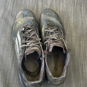 Svarta Elite Adidas fotbollsskor med dobbar - Säljer ett par svarta Adidas fotbollsskor Elite med silvriga ränder och subtila gulddetaljer. Skorna har snörning och en platt sula med fasta dobbar för gräsplan. Ovandelen är tillverkad i syntetmaterial med en sportig, strömlinjeformad design som ger grym känsla på planen.