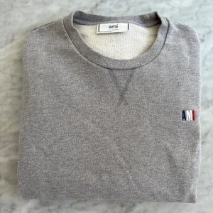Grå sweatshirt från AMI Paris - Snygg grå sweatshirt från AMI Paris med broderad logga i blått, vitt och rött på bröstet. Tröjan har rund hals, ribbade muddar och lång ärm. Perfekt för chill dagar och enkel att matcha! Tveka inte på att höra av dig vid frågor och funderingar 