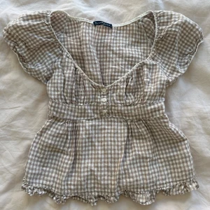 Rutig babydoll top från Brandy Melville - Supersöt rutig blus från Brandy Melville i beige och vitt. Blusen har korta puffärmar, v-ringning med spetskant, knappar framtill och volangdetaljer vid nederkanten. Tillverkad i mjuk bomull, perfekt för en chill och trendig look.