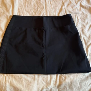 Svart kortkjol från Mango - Snygg svart kostym kortkjol från Mango Casualwear i klassisk rak modell. Kjolen har en diskret dragkedja bak och är gjord i ett mjukt polyestertyg som sitter bekvämt. Den är sönder vid dragkedjan men går lätt att sy ihop. Perfekt att matcha med en cool topp eller oversized jacka för en trendig look. Säljs då den är för liten 