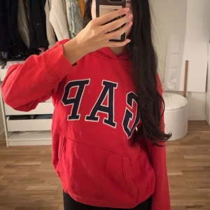 Röd hoodie från GAP  - Säljer en röd hoodie från GAP Kids med stor, broderad GAP-logga i marinblått och vitt på bröstet. Tröjan har huva, känguruficka och är tillverkad i mjuk bomullsmix. Perfekt för chill dagar eller när du vill ha en sportig look. Det är en storlek XL, passar dock tjejer som är runt 160/165cm som jag då det är barn storlek.😊