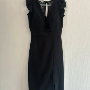 Svart långklänning med spets från Pedé - Snygg svart långklänning från Pedé med spetsdetaljer upptill och volang vid axlarna. Klänningen har en v-ringad front med dekorativ öppning och är ärmlös. Materialet är mjukt och har en elegant känsla, perfekt för dig som gillar stilrena och coola looks.