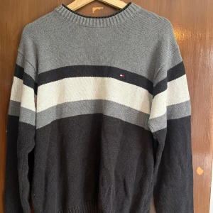 Randig stickad tröja Tommy Hilfiger - Snygg stickad tröja från Tommy Hilfiger i grå, svart och vit med breda ränder. Klassisk rund halsringning och diskret logga på bröstet. Perfekt för dig som gillar en stilren och avslappnad look.