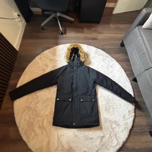 Svart parkasjacka från Everest XXS - Svart parkasjacka från Everest i storlek XXS. Jackan har avtagbar huva med fuskpäls, stora fickor framtill med knappar och dragkedja. Perfekt för kalla dagar med sin långa modell och varma foder. Snygg och funktionell vinterjacka med stilren look.