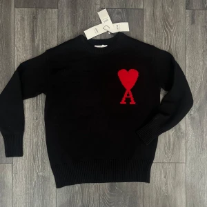 Svart stickad tröja från Ami Paris - Svart stickad tröja från Ami Paris med ikoniskt rött hjärta och A-logga på bröstet. Klassisk rund halsringning, ribbade muddar och långärmad modell. Perfekt för dig som vill ha en stilren men ändå unik look.