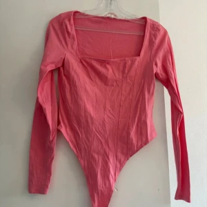Rosa långärmad bodysuit från H&M - Säljer en rosa långärmad bodysuit från H&M med fyrkantig urringning och markerade sömmar framtill. Bodyn har en tight passform och knäppning i grenen. Perfekt att styla med jeans eller kjol för en trendig look.
