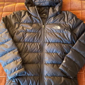 Svart dunjacka från The North Face - Säljer en svart dunjacka från The North Face med huva och dragkedja. Jackan är quiltad och har logga på bröstet samt 550-brodyr på ärmen. Perfekt för kalla dagar och riktigt skön att bära. Klassisk och stilren design som funkar till det mesta.