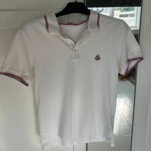 Vit Moncler piké med röd/blå detaljer - Vit pikétröja från Moncler med klassisk krage och korta ärmar. Tröjan har Moncler-logga broderad på bröstet och snygga röd/blå ränder på krage och ärmslut. Tillverkad i bomull för skön och luftig känsla. Perfekt för dig som gillar stilrena märkesplagg.