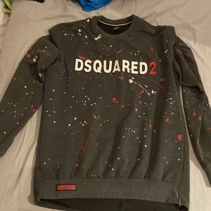 DSQUARED2 grå sweatshirt med färgstänk - Grå sweatshirt från DSQUARED2 i storlek L med coola färgstänk i vitt och rött över hela tröjan. Stor logga framtill och ribbade muddar. Skön passform och perfekt för dig som gillar streetstyle och statement-plagg.