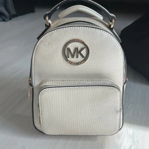 Beige ryggsäck från Michael Kors - Snygg beige ryggsäck från Michael Kors med guldiga detaljer och stort MK-logo framtill. Väskan har ett huvudfack med dragkedja, ett praktiskt ytterfack och justerbara axelremmar. Tillverkad i tåligt syntetmaterial med stilren design som passar till många outfits.