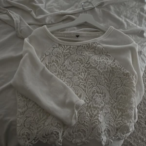 Vit croppad tröja med spetsdetaljer - Superfin vit croppad tröja med lång ärm och bred halsringning. Framsidan har detaljerad spets med blommönster och vågig kant nedtill. Perfekt att ha över ett linne 