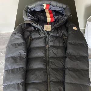 Mörkblå Moncler dunjacka med huva - Säljer en mörkblå dunjacka från Moncler med huva och snygga dragkedjor på bröstet. Jackan är quiltad och har en klassisk pufferstil som håller dig varm under kalla dagar. Perfekt för dig som vill ha en stilren och exklusiv vinterjacka.
