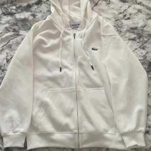 Vit hoodie från Lacoste med dragkedja - Snygg vit hoodie från Lacoste i regular fit med dragkedja framtill och klassisk krokodillogga på bröstet. Tröjan har huva med snörning, två fickor och långa ärmar. Perfekt för en clean och stilren look. Har aldrig används och den var för liten för mig när vi köpte den.