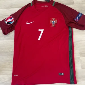 Portugal Ronaldo 7 Nike fotbollströja - Säljer en röd Portugal fotbollströja från Nike med nummer 7 och Ronaldo på ryggen. Tröjan har korta ärmar, gröna detaljer längs sidorna och officiella märken på ärmarna. Tillverkad i Dri-FIT material som andas, perfekt för match eller träning.