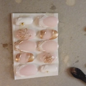 Rosa och guldiga lösnaglar med dekoration - Snygga lösnaglar i ljusrosa och vitt med detaljer i glittrande guld. Några naglar har blommor och andra har metallic-guldiga tippar och linjer. Setet innehåller olika former, mestadels oval, och små guldfärgade pärlor som dekoration. Perfekt för dig som vill ha extra glam på naglarna.