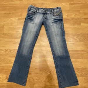 Lågmidjade Blå bootcut jeans från Divine - Snygga blå jeansbyxor från Divine med bootcut-snitt och broderade detaljer i gult på bakfickorna. Jeansen har slitningar framtill och klassisk femficksdesign. Perfekta för dig som gillar en avslappnad men ändå trendig look.