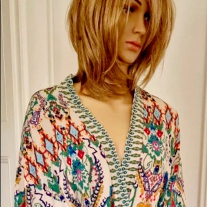 Mönstrad Positano-tunika kimono Zara med knyt - Färgstark och bohemisk positano-tunika kimono med vackert blommönster i grönt, blått, orange och rosa på ljus botten. Klänningen har omlott-look, knytband i midjan och långa ärmar med orange detalj. Perfekt för dig som gillar unika och färgglada plagg.