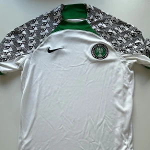 Nigeria vit fotbollströja från Nike Äkta - Snygg Nigeria fotbollströja från Nike i vit med gröna detaljer och svartvitt geometriskt mönster på axlarna. Kortärmad modell med rund hals och broderat landslagsemblem på bröstet. Perfekt för dig som vill sticka ut på planen eller på läktaren. Ny skick och jädrigt snygg. Köpt för 2000