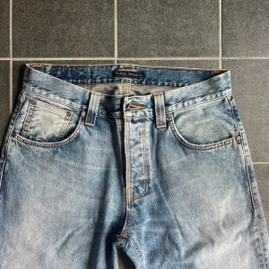 Nudie Jeans  - Säljer ett par ljusblå jeans från Nudie Jeans Co i klassisk femficksmodell. Jeansen har raka ben, orangea kontrastsömmar och snyggt slitna detaljer. Perfekta för dig som gillar en avslappnad och tidlös stil.